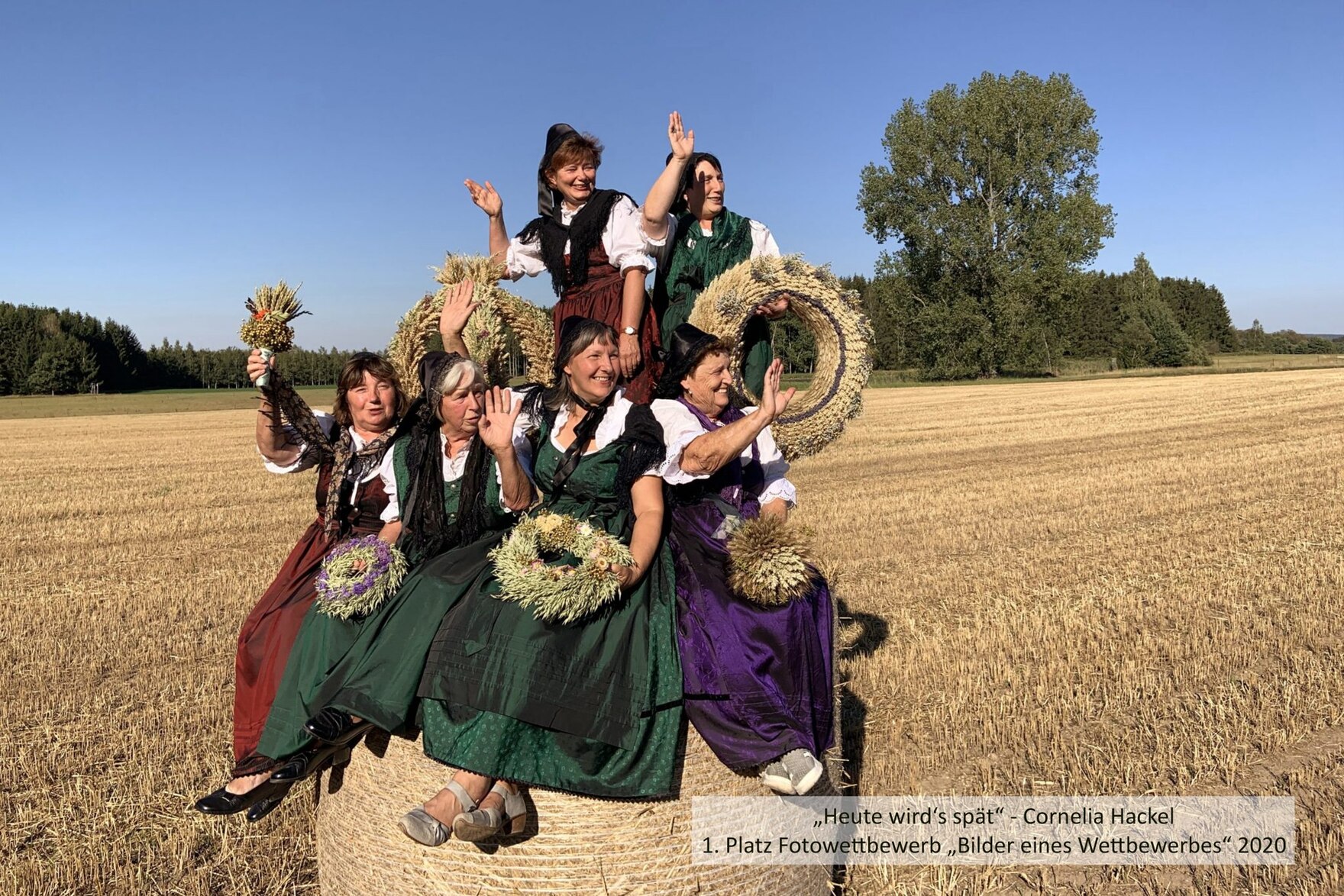 Landfrauen in Tracht sitzen auf einem Strohballen auf dem Feld, lachen und winken