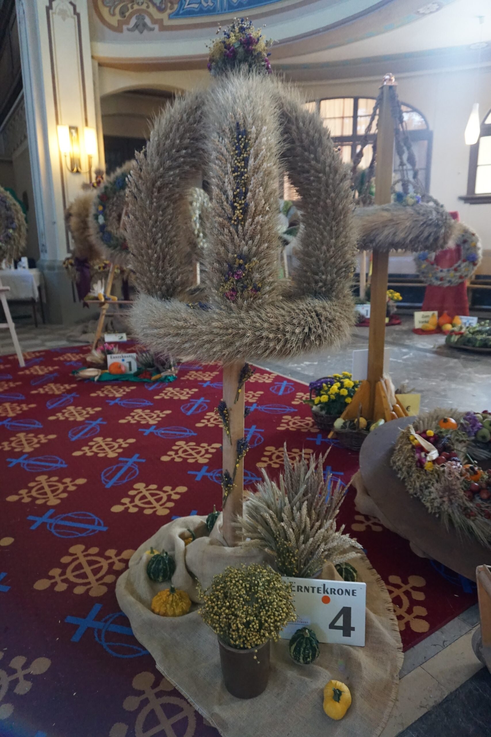 Erntekrone Nummer 4 aus Getreide mit Blumenschmuck und Dekoration aus Früchten und Blumen am Boden.