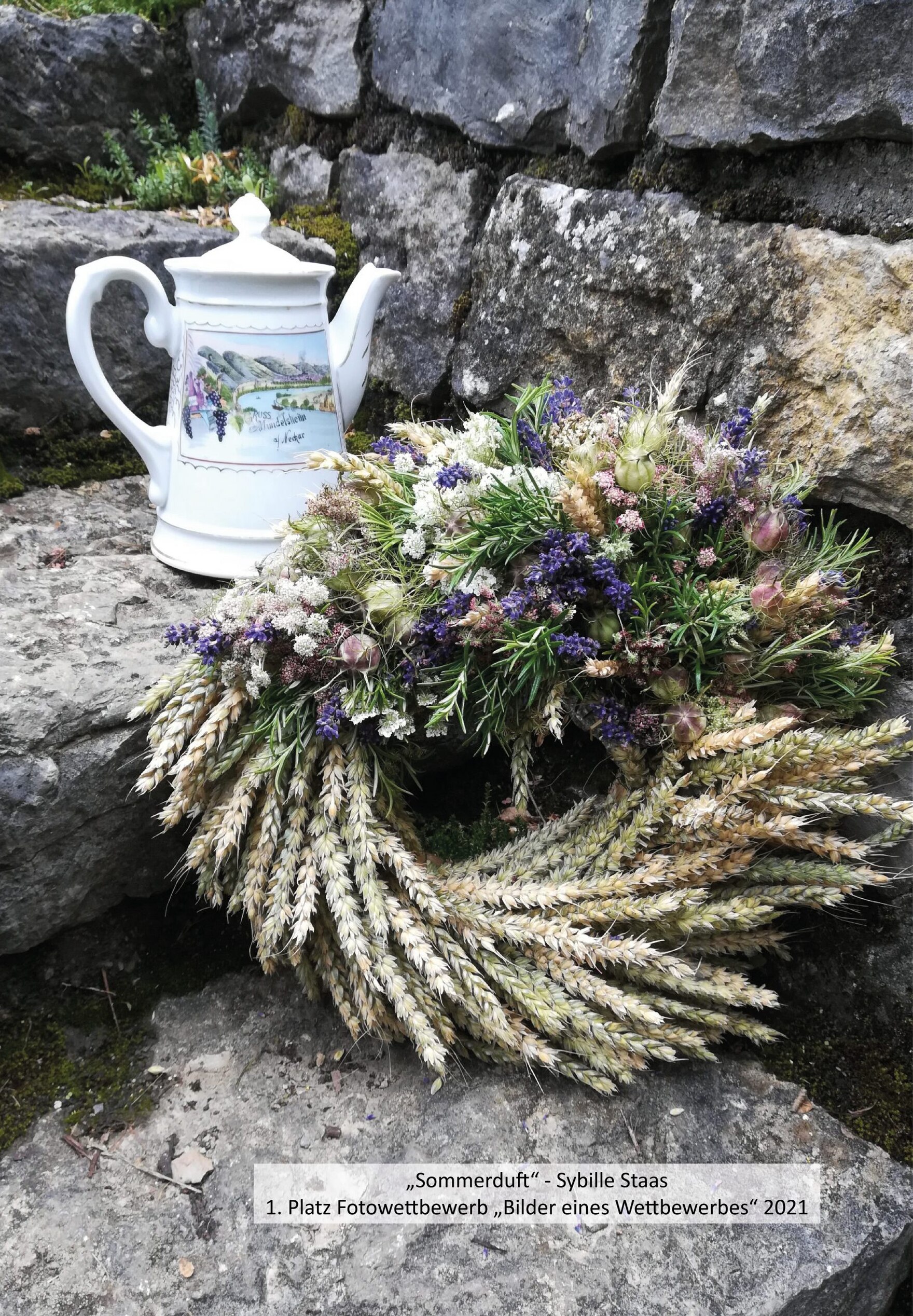 Auf grauen Treppenstufen vor einer Steinmauer liegt ein Getreidekranz mit Trockenblumen, dahinter steht eine Porzellankanne