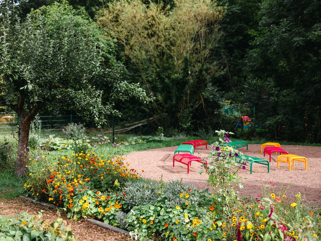 Bunte Bänke im Schulgarten der Grundschule Rödlitz bilden ein Klassenzimmer im Freien. Im Vordergrund Beete, ein Weg und ein Apfelbaum.