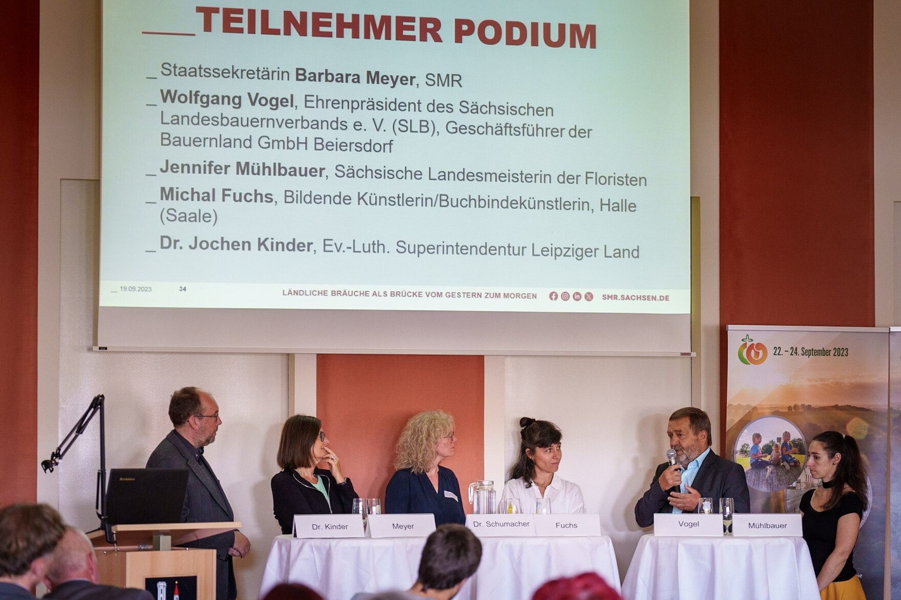 sechs Podiumsgäste stehen hinter Stehtischen, im Hintergrund sind die Namen eingeblendet