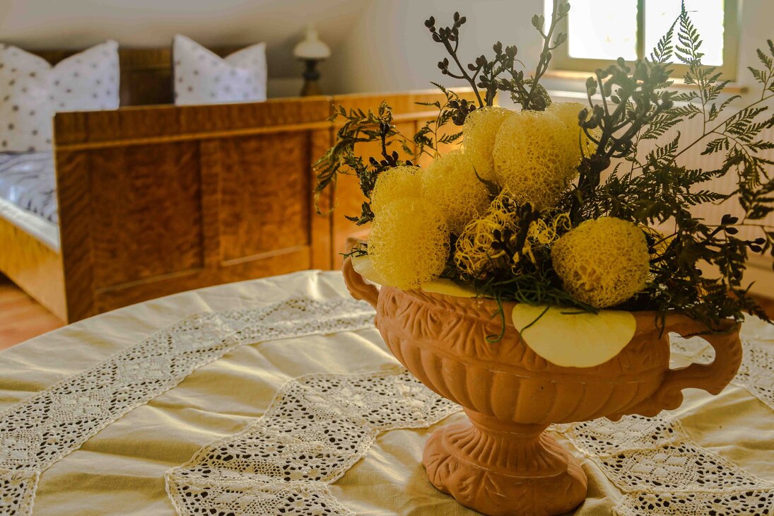 Auf gestickter Tischdecke stehendes Keramikgefäß mit Blumengesteck, im Hintergrund Doppelbett