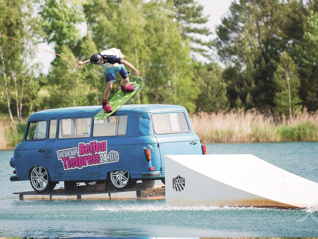 Sprungschanze im Wasser, die gerade ein Wassersportler auf einem Wakeboard absolviert hat, im Hintergrund ein blauer Barkas, der auf einer Schwimminsel steht